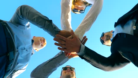 Young-business-partners-standing-and-stacked-hands-together-outdoor.-Successful-colleagues-putting-their-arms-in-centre-and-raising-them-up-to-blue-sky.-Unity-and-teamwork-concept.-Low-angle-view