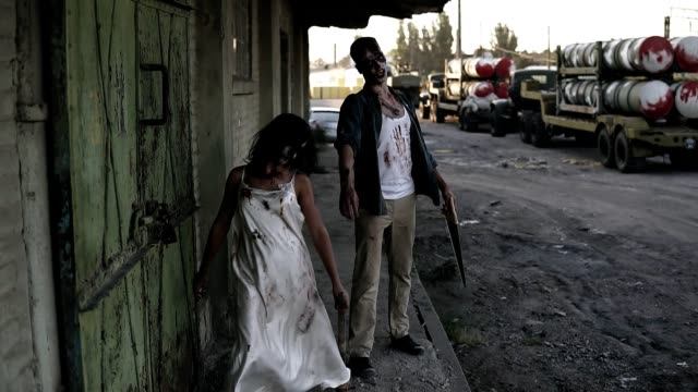 Portrait-of-creepy-male-and-female-ghost-or-zombie-walking-with-wounded-face-and-bloody-clothes.-Man-holding-saw-and-woman-is-with-a-stick.-Industrial,-abandoned-town-and-tracks-on-the-background