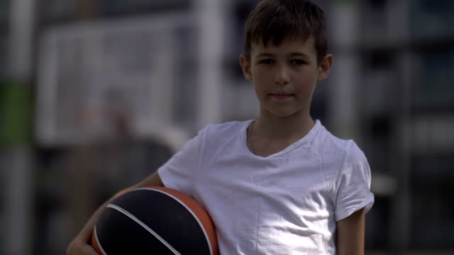 Retrato-de-un-niño-con-una-pelota-de-basket-en-una-cancha-de-baloncesto,-mira-a-la-cámara,-al-aire-libre