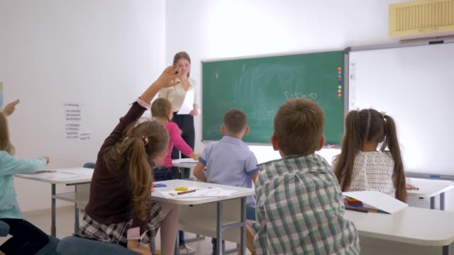 Lehrer-steht-vor-der-Klasse-in-der-Nähe-von-Blackboard-und-bittet-Schüler-Frage,-sie-ihre-Hände-heben,