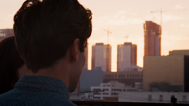 Rückansicht-des-Paares-auf-auf-dem-Dachterrasse-mit-Blick-über-Stadt-Skyline-bei-Sonnenuntergang