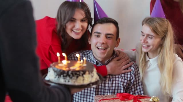 Handsome-man-blowing-birthday-candles-while-close-friends-throwing-a-surprise-party,-slowmotion-in-cozy-modern-apartment