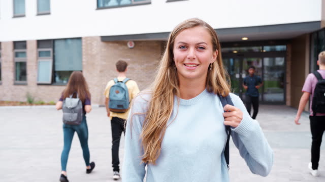 Portrait-Of-Smiling-Female-High-School-Student-Outside-College-Building-With-Other-Teenage-Students-In-Background