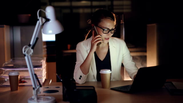 Cute-girl-company-manager-is-talking-on-mobile-phone-and-using-laptop-sitting-at-desk-in-dark-office-and-smiling-enjoying-her-job-and-holding-to-go-coffee.