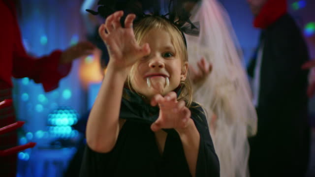 Halloween-Costume-Party:-Cute-Little-Bat-Girl-with-Sharp-Fangs-Makes-Scary-Funny-Faces.-In-the-Background-Monster-Party-in-Decorated-Room-and-Disco-Lights