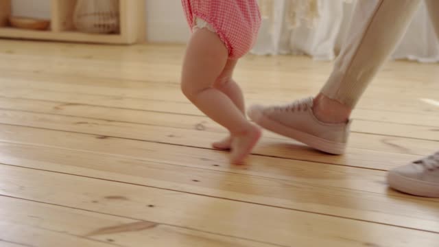 Tracking-close-up-shot-of-unrecognizable-caring-mother-helping-her-barefoot-baby-daughter-walking-towards-sofa-at-home
