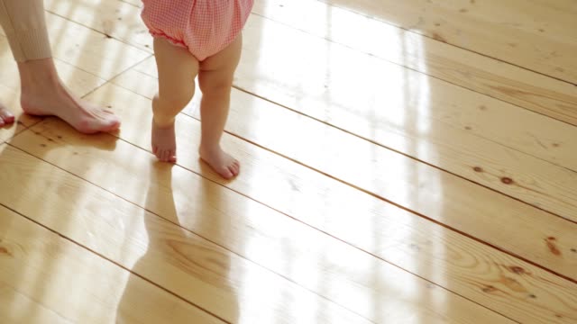 Liebenswerte-Baby-Mädchen-lernen,-auf-Hartholzboden-zu-Hause-zu-Fuß-von-ihrer-liebevollen-Mutter-unterstützt,-Tracking-erschossen