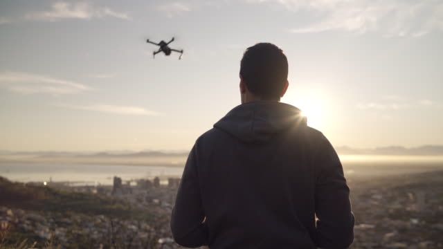 silhouette-shot-of-a-man-flying-his-drone-over-a-city-scape-during-sunrise-on-a-bright-sunny-day-outdoors---futuristic-technology-used-as-personal-hobby