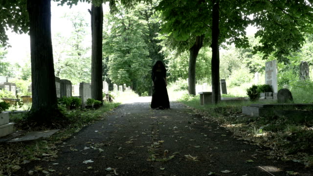 Woman-in-mourning-black-clothes-walking-slowly-on-alley-in-cemetery-holding-a-flower-crown-in-her-hand-portrait-of-sorrow-and-loneliness