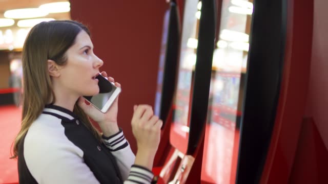 Teenager-buying-movie-ticket-from-vending-machine-at-cinema.-Woman-using-mobile-talking-to-friend