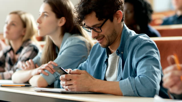 Hispanic-Mann-nutzt-Smartphone-während-einer-Vorlesung-im-Klassenzimmer.-Junge-helle-Studenten-studieren-an-der-Universität.