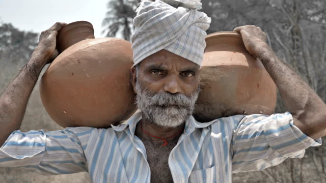 Tired-poor-middle-aged-man-carrying-two-clay-pots-standing-in-dry-farmland