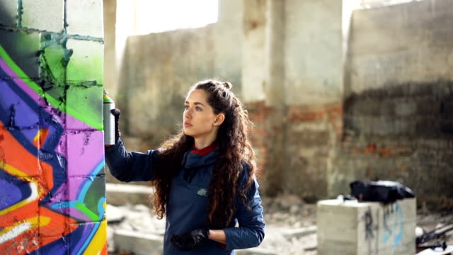 Slow-motion-of-graffiti-artist-painting-on-wall-in-abandoned-building-using-aerosol-spray-paint.-Attractive-girl-with-curly-hair-is-busy-with-her-work,-she-is-looking-at-painting.