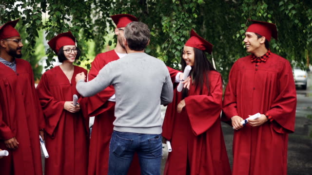 Slow-motion-of-proud-university-teacher-hugging-his-students-and-shaking-hands,-graduating-students-are-standing-in-line-with-diplomas-and-laughing.
