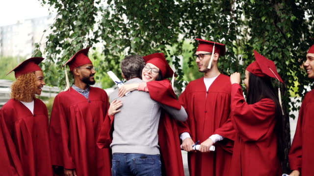 Slow-motion-of-college-teacher-hugging-his-students-and-shaking-hands-on-graduation-day-with-joy-and-pride.-Green-trees,-educational-institution-building-is-visible.