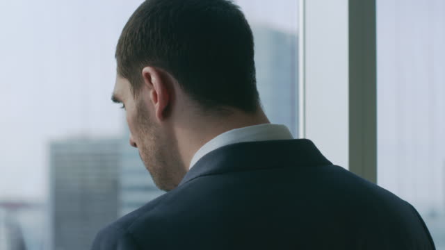 Close-up-Shot-of-the-Confident-Businessman-in-a-Suit-Standing-in-His-Office-and-Making-Phone-Call-so-He-can-Close-the-Deal,-Looking-out-of-the-Window.-Big-City-Business-District-Panoramic-View.