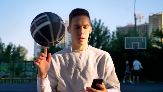 Handsome-man-spinning-basketball-on-finger-and-taking-selfie-or-having-video-chat,-wearing-earphones,-smiling,-park-in-background