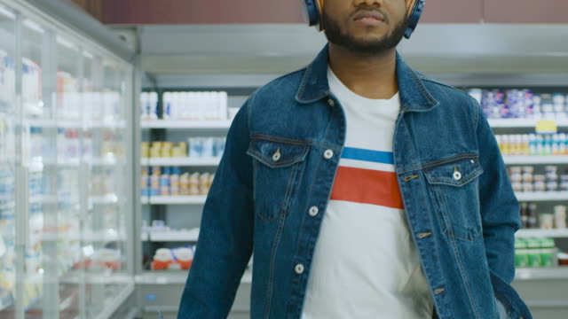 At-the-Supermarket:-Stylish-African-American-Guy-with-Headphones-Walks-Through-Frozen-Goods-Section-of-the-Store.-Following-Back-View-Shot.-Slow-Motion.