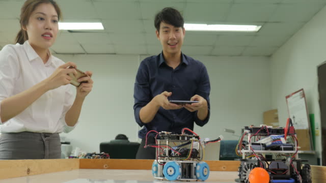 Two-electronics-engineer-programming-and-control-robots-to-playing-soccer-with-mobile-phone-in-the-workshop.-People-with-technology-or-innovation-concept.
