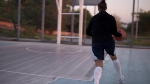Backside-footage-of-a-young-girl-basketball-player-training-and-exercising-outdoors-on-the-local-court.-Dribbling-with-the-ball,-bouncing-and-make-a-shot