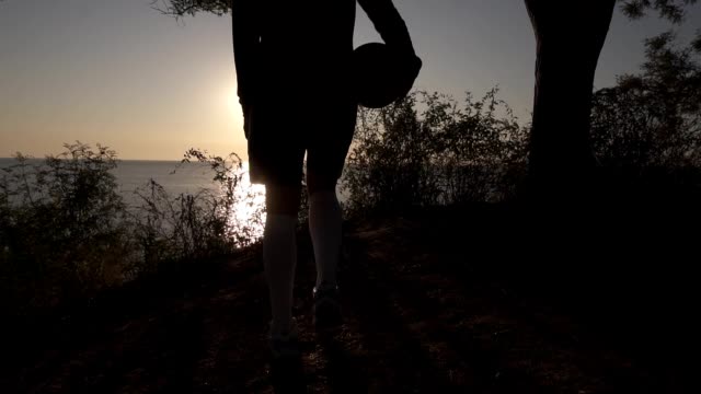A-silhouette-of-a-basketball-player-girl-comes-with-a-ball-in-her-hand,-coming-up-to-the-slope-with-trees-around.-Looks-at-the-sun-shining-over-the-sea.-Morning-dusk.-Backside-view
