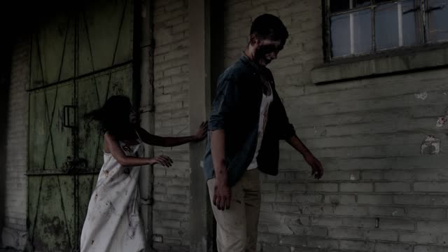 Two-zombies-are-walking-with-an-abandoned-house-on-the-background.-Brunette-girl-with-wounded-face-and-bloody-white-dress-and-wounded-male-zombie-are-walking-outdoors.-Accelerating-footage