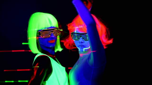 Women-with-laser,-UV-face-paint,-wig,-glowing-glasses,-glowing-clothing-dancing-together-fast-in-front-of-camera,-Half-body-shot.-Caucasian-and-asian-woman.-.