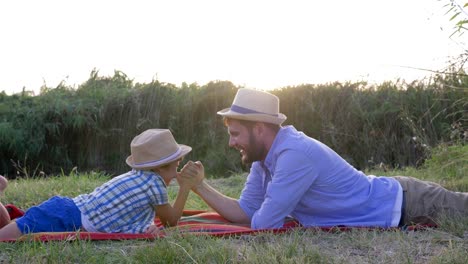 loving-father-and-kid-play-together-in-arm-wrestling-lying-on-plaid-during-weekend-leisure-outdoors