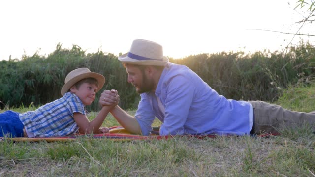 happy-father-and-son-kid-play-together-in-arm-wrestling-lying-on-plaid-outdoors-on-background-of-sunset-in-rural