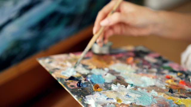 Close-up-shot-of-dirty-female-hand-holding-paintbrush-and-mixing-colors-on-palette-then-painting-marine-landscape-on-canvas.-Art-and-tools-concept.