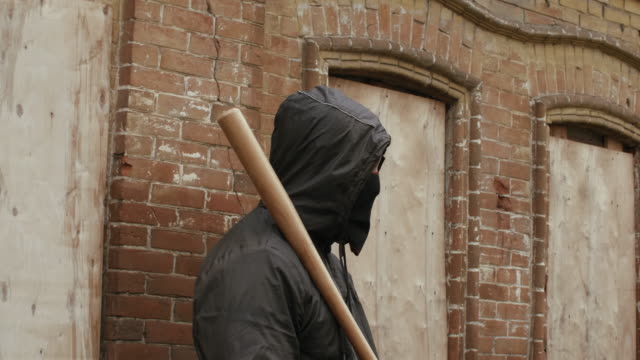 Street-hooligan-in-black-mask-with-baseball-bat-on-criminal-street-district-looking-to-camera