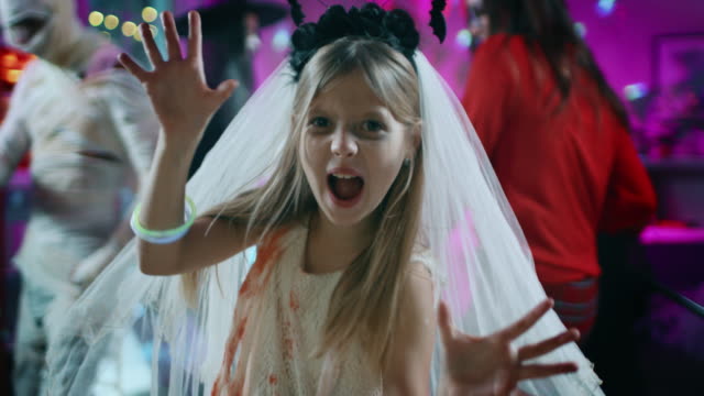 Halloween-Kostümparty:-Little-Girl-in-a-Bloody-White-Bride-Dress-dreht-sich-um-und-macht-Scary-Faces.-In-der-Hintergrundegruppe-der-Monster-tanzen-und-Spaß-haben-im-dekorierten-Raum-mit-Disco-Lights