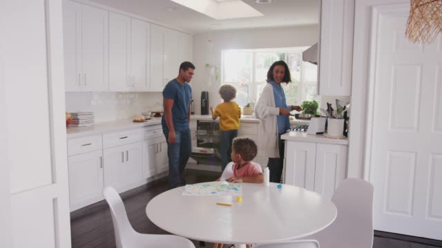 Papá-viendo-hijo-niño-pintando-en-la-cocina,-madre-e-hija-preparan-la-comida-en-el-fondo