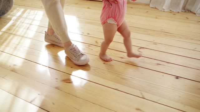 Tracking-right-shot-of-mother-supporting-her-barefoot-baby-daughter-in-pink-bodysuit-learning-to-walk-on-hardwood-floor-at-home