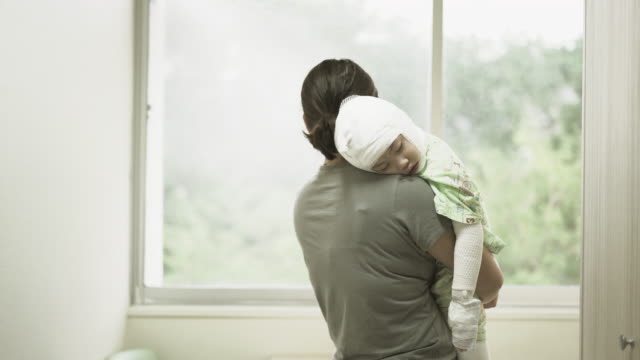 Asian-mother-was-holding-a-sick-baby-girl-at-her-shoulder-beside-the-window-in-hospital,-Girl-are-sick-with-Atopic-dermatitis.-She-was-wrapped-with-a-bandage-on-the-head-neck-arms-and-legs.-Real-live