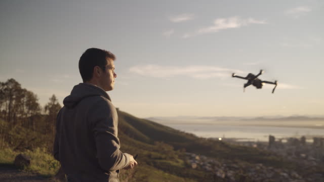 Gimbal-Aufnahme-eines-Mannes,-der-seine-Drohne-steuert,-so-dass-sie-während-des-Sonnenaufgangs-über-die-Stadt-fliegen---sehr-filmischer-Clip-mit-Sonnenfackeln