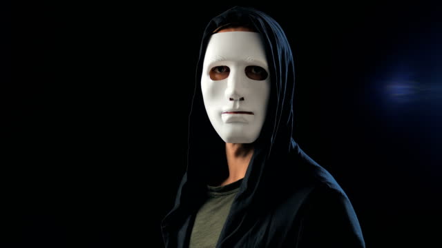 Ein-anonymer-Mann-in-einer-weißen-Maske,-die-sein-Gesicht-bedeckt,-und-eine-schwarze-Kapuze-dreht-sich-und-schaut-in-die-Kamera.-Schwarzer-dunkler-Hintergrund.-Konzept-eines-Mitglieds-einer-geheimen-Organisation