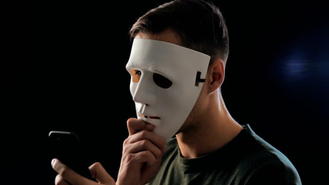 Betrüger-verstecken-sein-Gesicht-mit-Mast-und-fordert-ein-Lösegeld-für-Erpressung-mit-einem-Smartphone.-Maskierter-Krimineller-schüchtert-das-Opfer-mit-Hilfe-von-Drohungen-per-SMS-per-Handy-ein.