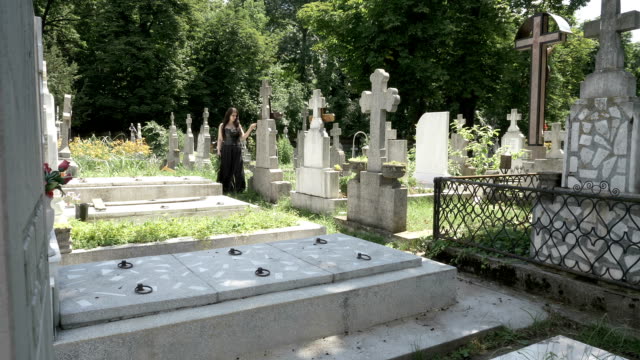 Chica-gótica-fúnebre-caminando-al-lado-de-lápidas-en-cementerio-antiguo-contemplando-la-muerte-y-la-soledad