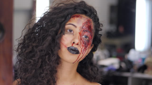 Scary-portrait-of-young-girl-with-Halloween-blood-makeup.-Beautiful-latin-woman-with-curly-hair-looking-into-mirror-reflection-in-the-dressing-room.-Preparation-for-celebrating.-Slow-motion