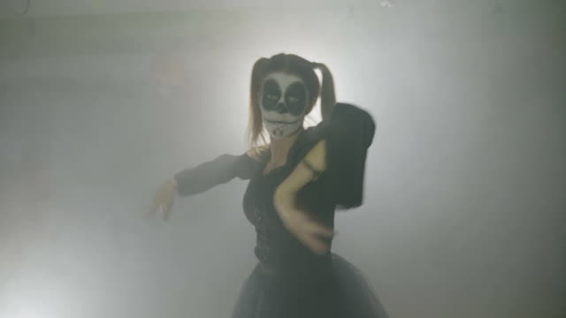 Young-female-teenager-dressed-as-a-sinister-doll-exercising-her-zombie-dancing-moves-in-fog-for-halloween-night-at-club