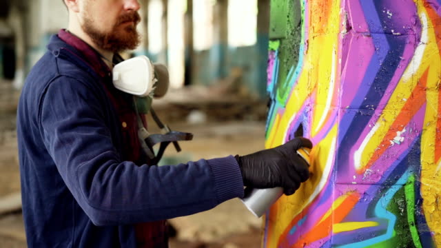 Bärtiger-Mann-Graffiti-Maler-nutzt-Aerosol-Farbe-um-Säule-im-alten-Industriegebäude-zu-schmücken.-Moderne-Urbane-Kunst,-junge-kreative-und-Hobby-Konzept.