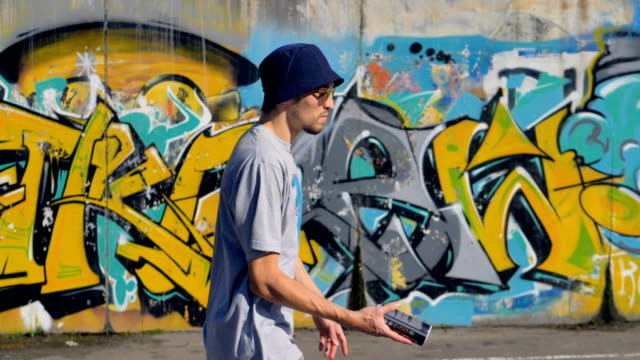 Young-man-is-walking-along-the-street-with-graffiti-painting-on-the-background-and-a-spray-in-his-hands