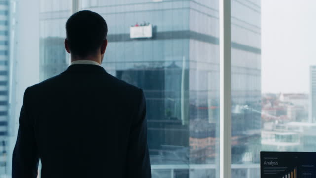 Following-Shot-of-the-Confident-Businessman-in-a-Suit-Walking-Through-His-Office-and-Looking-out-of-the-Window-Thoughtfully.-Stylish-Modern-Business-Office-with-Personal-Computer-and-Big-City-View.