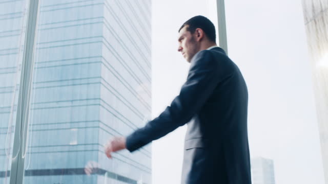 Low-Angle-Following-Shot-of-the-Confident-Businessman-in-a-Suit-Walking-Through-His-Office-and-Looking-out-of-the-Window-Thoughtfully.-Stylish-Modern-Business-Office-with-Big-City-View.