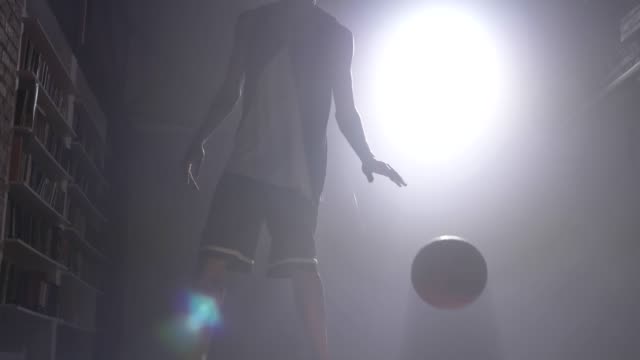Silueta-de-jugador-de-uno-baloncesto-jugando-con-bola-en-habitación-oscura-niebla-con-proyector