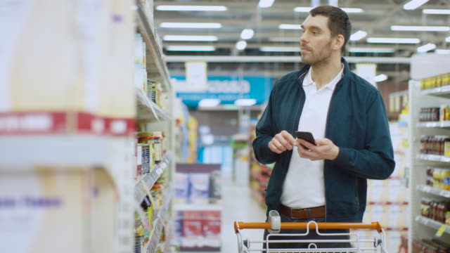 Im-Supermarkt:-gut-aussehender-Mann-nutzt-Smartphone-und-nimmt-Bild-der-Dose-waren.-Er-steht-mit-Einkaufswagen-in-Konserven-Abschnitt.