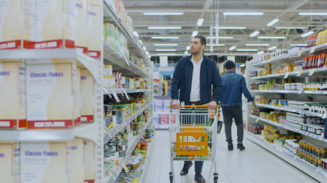 At-the-Supermarket:-Handsome-Man-Browses-Through-Shelf-with-Canned-Goods,-Looks-at-Tin-Can-but-Decided-not-to-Buy-it.-He-Proceeds-Walking-with-Shopping-Cart-Through-Different-Sections-of-the-Store.