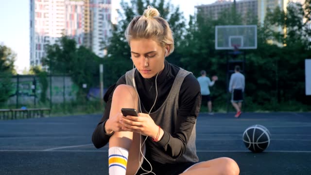 Pensive-young-beautiful-woman-sitting-on-basketball-court-and-typing-on-phone,-wearing-earphones,-men-playing-in-background