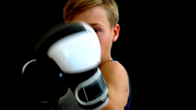 A-teenager-in-boxing-gloves-stands-on-a-dark-background.-The-boy-has-blond-hair,-a-gray-T-shirt.-White-black-gloves-almost-cover-his-face